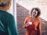 Photo focused on a smiling woman holding up a speakerphone with one hand. In the background there are other people, smiling, but blurred slightly.