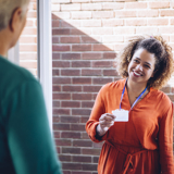 Photo focused on a smiling woman holding up a speakerphone with one hand. In the background there are other people, smiling, but blurred slightly.