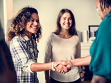 Two women meeting another woman (who has her back to the viewer), shaking hands and smiling.